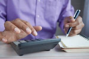 ROAS versus ROI: homem negro com camisa roxa usando calculadora e bloco de notas com caneta na mão.
